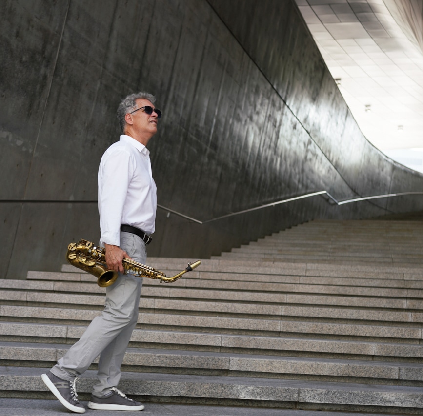 Jim Snidero stands on a concrete staircase, holding his sax. He wears sunglasses and looks up to the sky, side-on to the viewer.