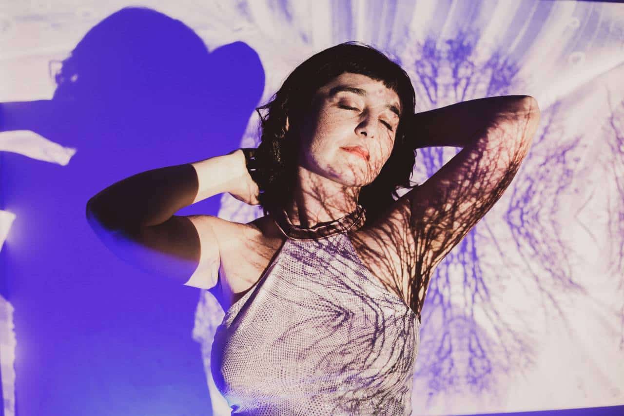 Camila Meza stands with her arms raised and eyes closed, with images of tree branches projected onto her body.